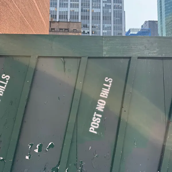 One-line white POST NO BILLS stencil set diagonally across hunter-green scaffold plywood, with Midtown buildings above.