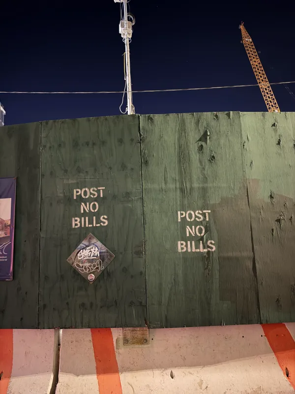 A green plywood construction fence at night, stenciled twice in white with "POST NO BILLS," with a graffiti-tagged diamond sticker below one of the stencils. Orange-and-white concrete barriers line the bottom, and a light pole and a yellow crane stand against the dark sky above.