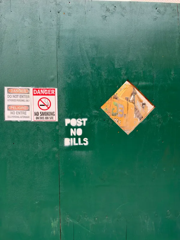 A dark green plywood construction wall with a white stenciled "POST NO BILLS" in the middle. To the left are posted signs reading "DANGER DO NOT ENTER AUTHORIZED PERSONNEL ONLY / PELIGRO NO ENTRE SOLO PERSONAL AUTORIZADO" and "DANGER NO SMOKING ON THIS JOB SITE," and to the right a diamond-shaped cutout shows a yellow excavator behind the wall.