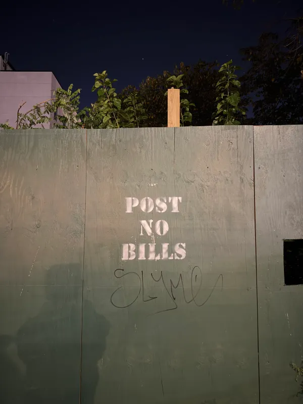 A green-painted plywood construction fence photographed at night, stenciled in white with "POST NO BILLS" and a black marker graffiti tag underneath. Leafy trees, a wooden post and the corner of a pale building rise above the fence against a dark sky.