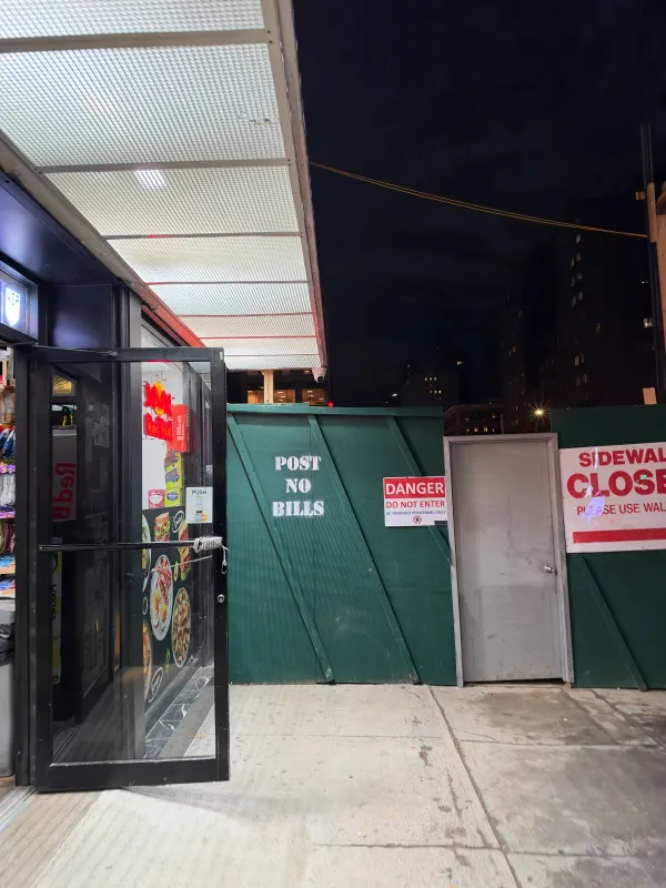 A green plywood construction fence at night has "POST NO BILLS" stenciled on it in white, next to a red "DANGER DO NOT ENTER" sign, a gray metal door, and a partly visible "SIDEWALK CLOSED" sign. On the left, a lit deli's glass door stands propped open under a bright awning.
